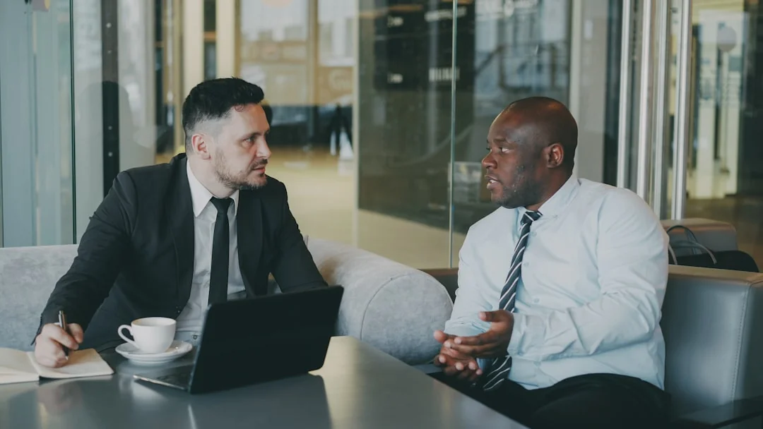 Two businessmen analyze stock market data using a smartphone and laptop in a modern office setting.